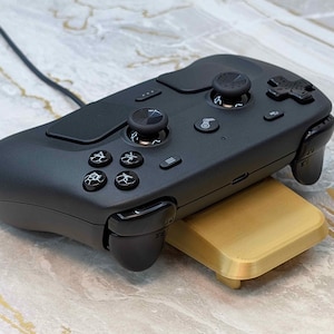May include: A black gaming controller with multiple buttons, joysticks, and a directional pad. The controller is resting on a gold-colored stand. A black cable extends from the top of the controller.