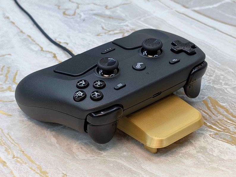 May include: A black gaming controller with multiple buttons, joysticks, and a directional pad. The controller is resting on a gold-colored stand. A black cable extends from the top of the controller.