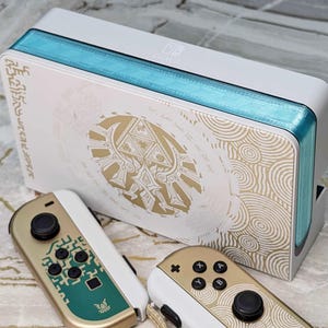 May include: A white Nintendo Switch console with gold and teal accents, featuring a gold design and matching controllers. The console has a teal stripe along the top and sides. The controllers are gold and white.