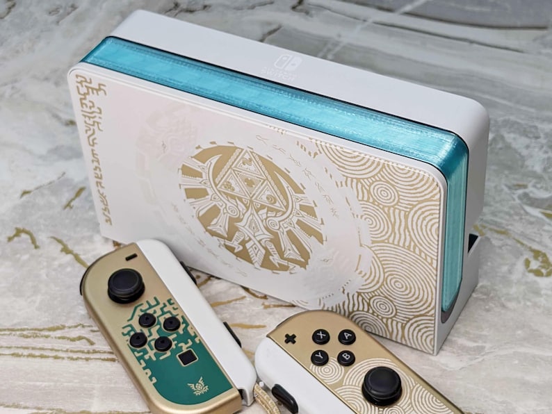 May include: A white Nintendo Switch console with gold and teal accents, featuring a gold design and matching controllers. The console has a teal stripe along the top and sides. The controllers are gold and white.