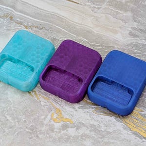 May include: Three rectangular, open-top containers in shades of blue, purple, and teal. Each has a textured honeycomb pattern on the top and a rectangular cutout on one side. The containers are arranged on a marbled surface.