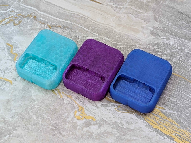 May include: Three rectangular, open-top containers in shades of blue, purple, and teal. Each has a textured honeycomb pattern on the top and a rectangular cutout on one side. The containers are arranged on a marbled surface.