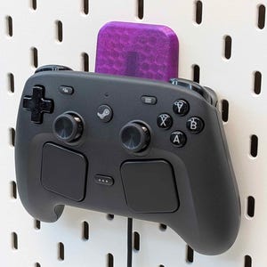 May include: A black Steam Deck controller is mounted on a white pegboard with a purple holder. The controller has various buttons, joysticks, and a D-pad. The pegboard has evenly spaced holes.