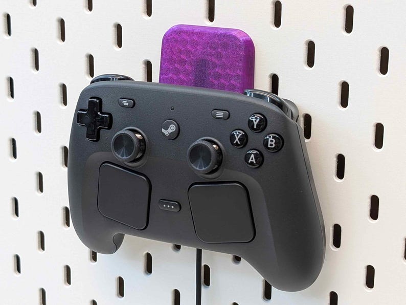 May include: A black Steam Deck controller is mounted on a white pegboard with a purple holder. The controller has various buttons, joysticks, and a D-pad. The pegboard has evenly spaced holes.