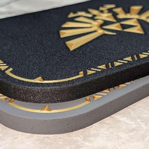 A closeup photograph of a Zelda style dock cover / faceplate for the Nintendo Switch 2, based on designs from the Legend of Zelda series. This closeup photo shows the difference between the Castle Gray finish and galaxy black glitter finish with silky gold colored highlights.