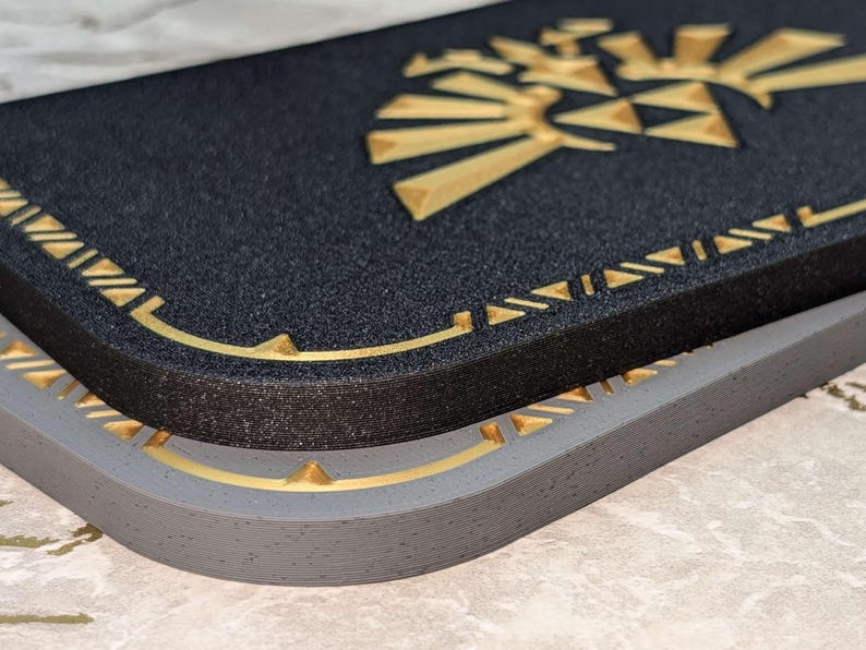 A closeup photograph of a Zelda style dock cover / faceplate for the Nintendo Switch 2, based on designs from the Legend of Zelda series. This closeup photo shows the difference between the Castle Gray finish and galaxy black glitter finish with silky gold colored highlights.