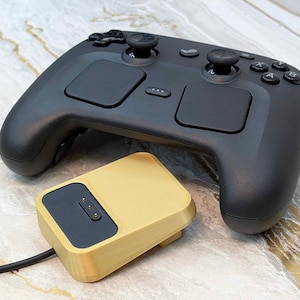 May include: A black gaming controller with two touchpads, two analog sticks, and several buttons. A gold-colored charging dock is connected to the controller with a black cable. The items are on a marble-patterned surface.