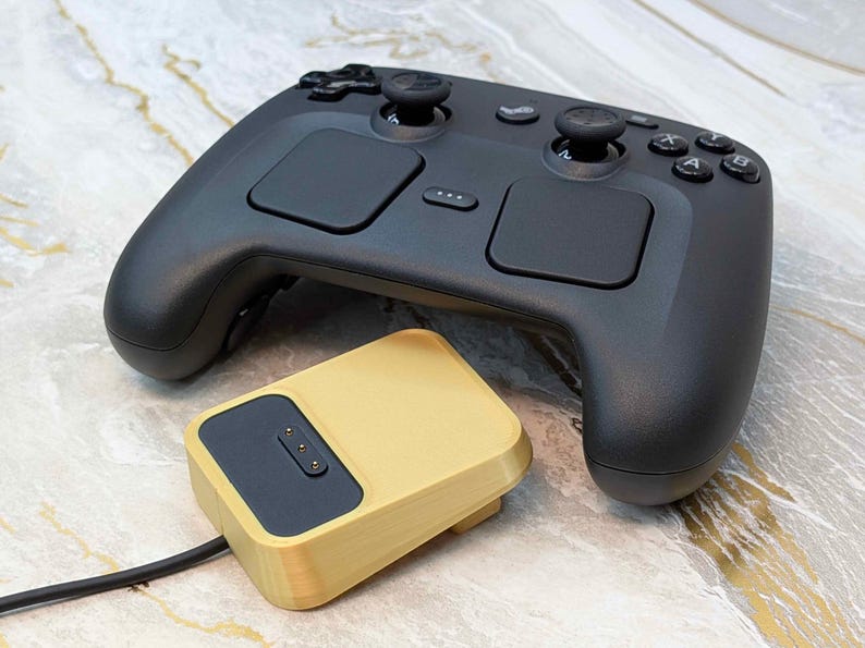 May include: A black gaming controller with two touchpads, two analog sticks, and several buttons. A gold-colored charging dock is connected to the controller with a black cable. The items are on a marble-patterned surface.