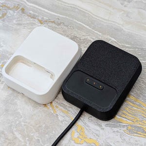 May include: Two rectangular charging docks. One is white with a recessed area, the other is black with a textured surface and three gold-colored charging pins. A black cable extends from the black dock.