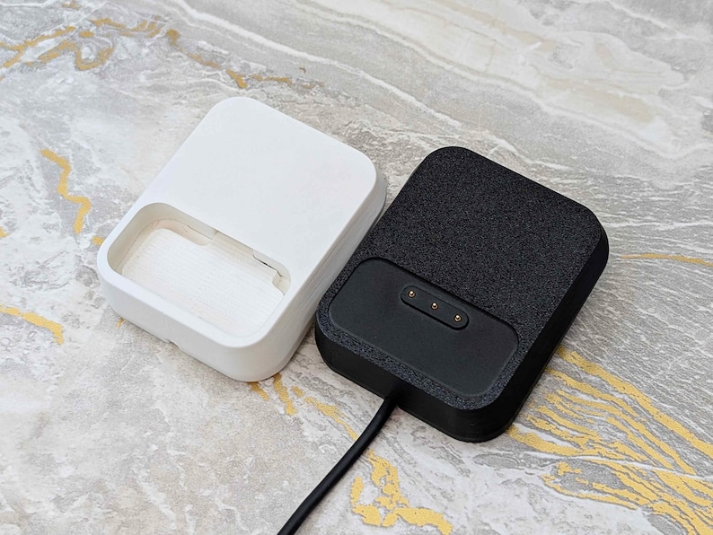 May include: Two rectangular charging docks. One is white with a recessed area, the other is black with a textured surface and three gold-colored charging pins. A black cable extends from the black dock.