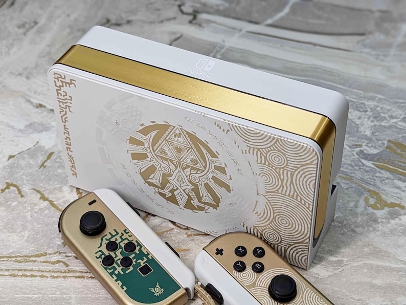 May include: White and gold Nintendo Switch docking station and Joy-Con controllers. The docking station features gold accents and a gold and white design with intricate patterns. The Joy-Con controllers are gold and white with black buttons and green accents.