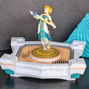 Może przedstawiać: Biała i złota platforma z niebiesko-zielonym wzorem spiralnym. Na platformie stoi figurka kobiety z blond włosami trzymającej miecz. Platforma ma okrągły kształt z złotym symbolem w środku.