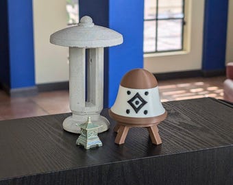 May include: Three decorative ceramic figurines on a black surface. A tall, white, cylindrical lantern with a roof, a small, green, pagoda-style lantern, and a brown and white bell-shaped figurine with a geometric design and four legs.