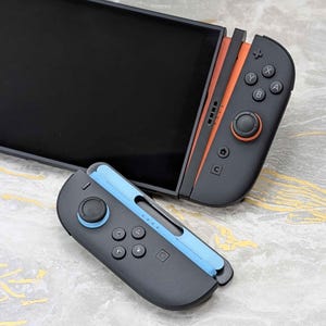 以下が含まれることがあります： 黒色の携帯ゲーム機で、オレンジと青のアクセントが付いています。コンソールには黒い画面と、オレンジ色のアクセントが付いたコントローラーと青色のアクセントが付いたコントローラーの2つの取り外し可能なコントローラーがあります。コントローラーにはボタンとジョイスティックがあります。