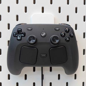 May include: A black video game controller with a Steam logo, mounted on a white pegboard. The controller has two analog sticks, a directional pad, and several buttons. The pegboard has evenly spaced holes.