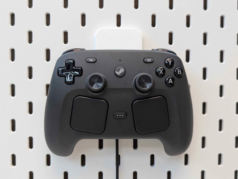 May include: A black video game controller with a Steam logo, mounted on a white pegboard. The controller has two analog sticks, a directional pad, and several buttons. The pegboard has evenly spaced holes.