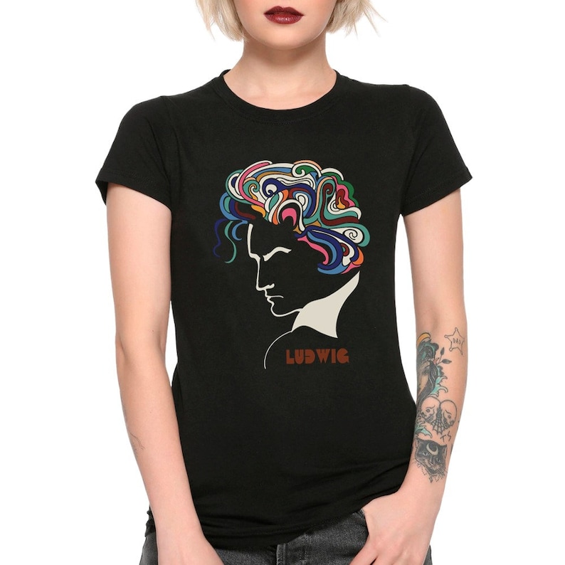Op de afbeelding: Zwart t-shirt met een profiel van een persoon met kleurrijk, abstract haar. Het woord "LUDWIG" is in bruin gedrukt. Een grafisch en artistiek ontwerp.