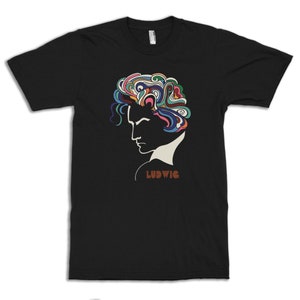 Op de afbeelding: Zwart T-shirt met een profiel van een persoon met kleurrijk, wervelend haar. Het ontwerp bevat de naam "LUDWIG" in bruine letters. Het shirt heeft een ronde hals.