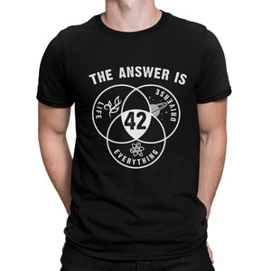 Può includere: T-shirt nera con un design grafico bianco. Il design presenta tre cerchi sovrapposti con le parole "Life", "Universe" e "Everything". Il numero "42" è al centro. Il testo "THE ANSWER IS" è sopra i cerchi.