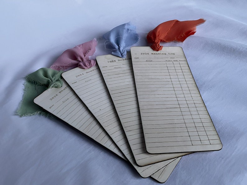 4 unique wooden library list bookmarks laying on top of one another. Each has a different colored ribbon tied to the top and it shows how the file looks like in real life. Each one had been designed on Canva and was cut & engraved by a laser engraver