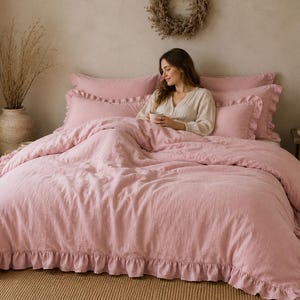 May include: A dusty pink bedding set featuring a duvet cover and matching pillowcases with ruffled edges. The fabric has a visible textured weave, and the set includes multiple pillows arranged against the headboard, creating a layered look.