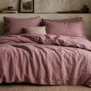 May include: A dusty rose linen duvet cover set featuring a row of visible buttons along the bottom edge. The bedding includes two matching pillowcases and a single rectangular accent pillow in a neutral beige tone, all arranged on a bed.