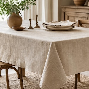 May include: A rectangular beige linen tablecloth with a visible woven texture draped over a dining table. The fabric features a natural, light tan color and a simple hemmed edge, providing a neutral foundation for the table setting.