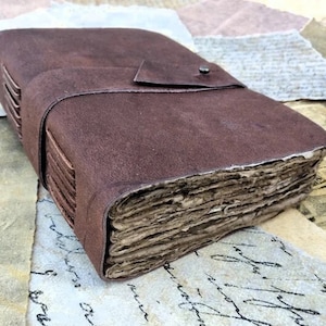 May include: A brown leather-bound journal with a flap closure and a metal button. The journal has a rustic, aged look and is made of thick, textured paper.