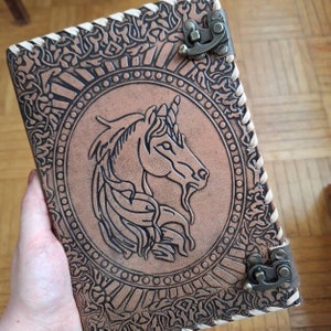 May include: A brown leather journal with a detailed embossed unicorn design on the cover. The journal features a decorative border, a metal clasp, and stitched edges. The cover is a light brown color.