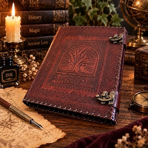 May include: A dark red leather-bound journal featuring an embossed tree design on the front cover, intricate border patterns, and two antique brass-colored metal latches. The journal has visible stitching along the edges and a rectangular central panel.