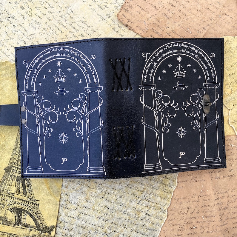 May include: Black leather journal with a white design of a gate with trees and stars. The text "mab cldd hoog ded clybage add dad ahogy ilang adtagbad qad beambla" is written around the gate.