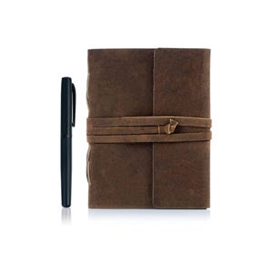 Puede incluir: Un diario de cuero marr&oacute;n con cierre de correa de cuero. El diario est&aacute; cerrado y tiene un bol&iacute;grafo a su lado.