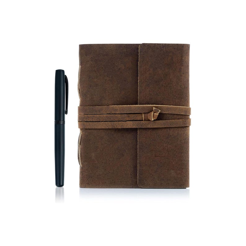 Puede incluir: Un diario de cuero marr&oacute;n con cierre de correa de cuero. El diario est&aacute; cerrado y tiene un bol&iacute;grafo a su lado.