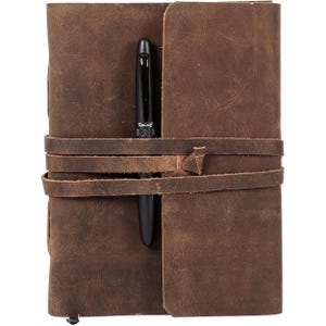 Puede incluir: Un diario de cuero marr&oacute;n con un bol&iacute;grafo metido en la correa de cuero. El diario tiene un aspecto desgastado y est&aacute; cerrado.