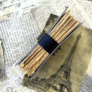 May include: A vintage leather-bound journal with a black leather strap closure. The journal is open to reveal pages made of thin, aged paper. The journal is lying on top of a vintage postcard featuring a black and white image of the Eiffel Tower.