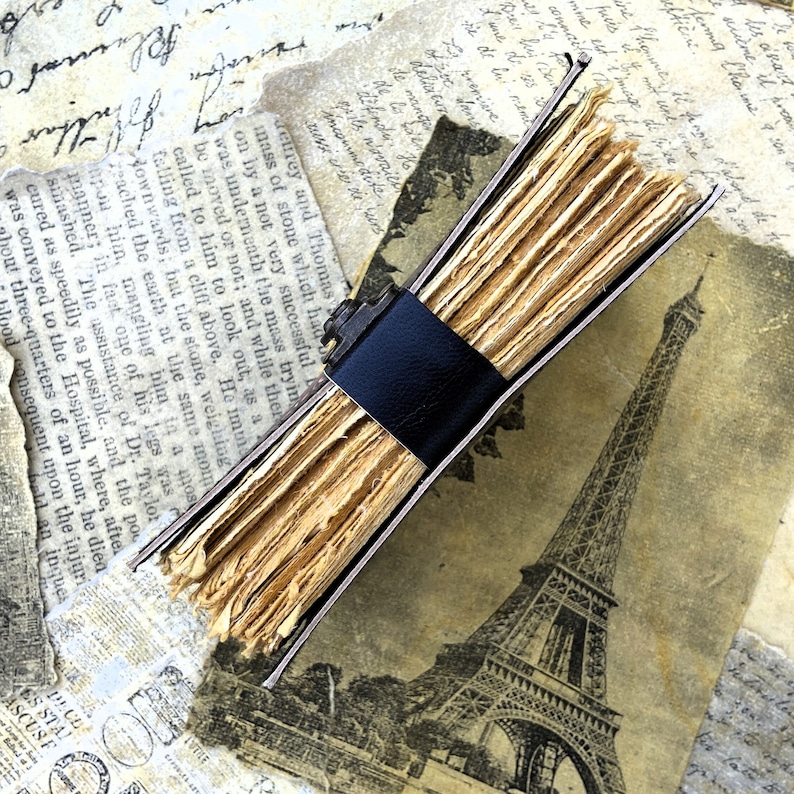 May include: A vintage leather-bound journal with a black leather strap closure. The journal is open to reveal pages made of thin, aged paper. The journal is lying on top of a vintage postcard featuring a black and white image of the Eiffel Tower.