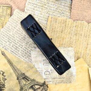 May include: A black leather journal with a decorative stitched design. The journal is lying on a pile of old, yellowed paper.