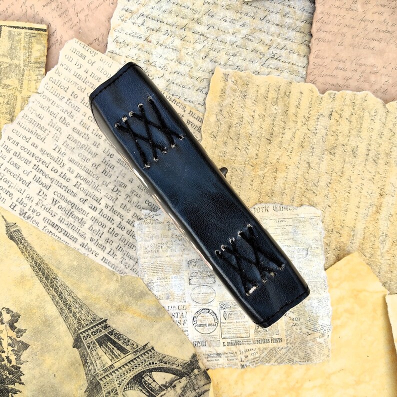 May include: A black leather journal with a decorative stitched design. The journal is lying on a pile of old, yellowed paper.