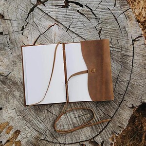 Puede incluir: Un diario de cuero marr&oacute;n con una p&aacute;gina blanca en blanco abierta. El diario se cierra con una correa de cuero marr&oacute;n y tiene una correa de cuero marr&oacute;n con una hebilla.