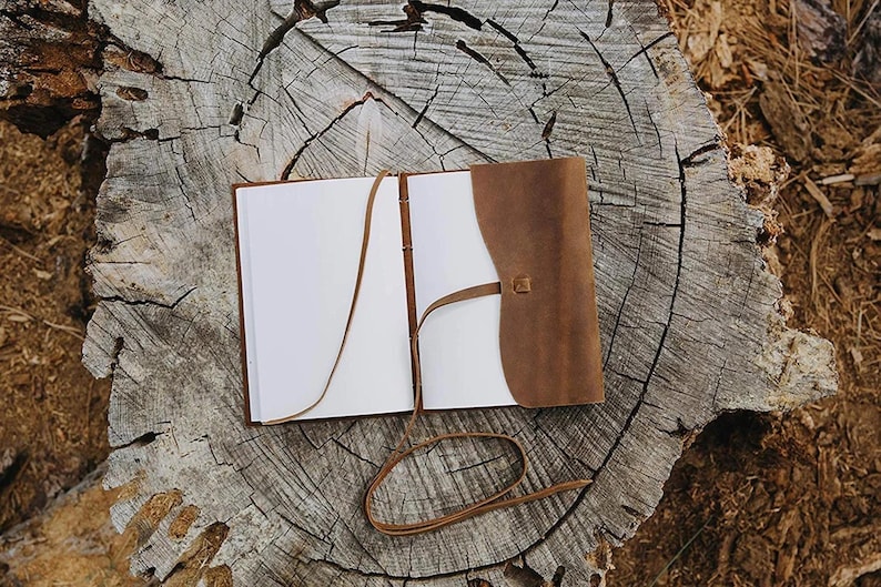 Puede incluir: Un diario de cuero marr&oacute;n con una p&aacute;gina blanca en blanco abierta. El diario se cierra con una correa de cuero marr&oacute;n y tiene una correa de cuero marr&oacute;n con una hebilla.