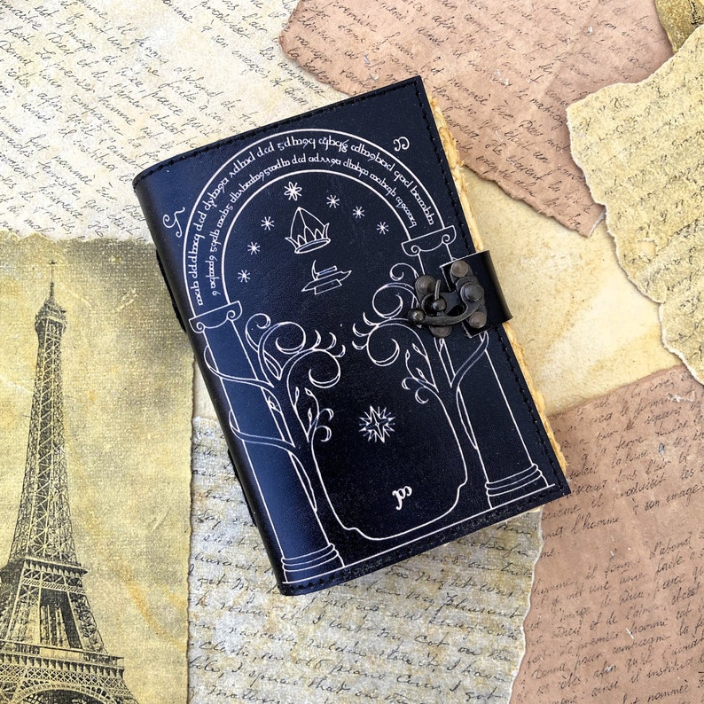 May include: Black leather journal with a white design of a tree with a door and stars. The journal has a metal clasp and the text "ab cld deg ded dynga adhaedded 5da99 sangg adbasbod god bombba" is written on the cover.