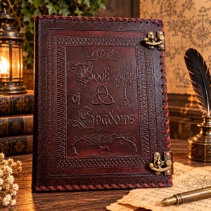 May include: A dark brown leather-bound journal featuring the embossed text Book of Shadows, a triquetra symbol, a tree, and a pentagram. The cover is secured with two antique brass metal latches and features decorative red leather stitching along the edges.