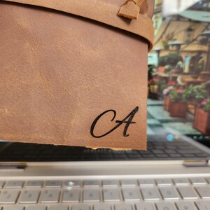 Puede incluir: Un diario de cuero marr&oacute;n con una correa de cuero negro y la letra "A" grabada en la parte delantera.