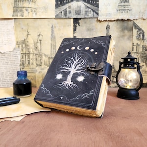 Peut inclure: Un journal en cuir noir avec un motif d'arbre et de lune blanc. Le journal a un fermoir et est ouvert pour révéler des pages vierges.