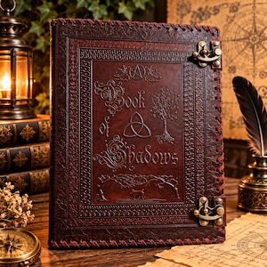 Może przedstawiać: Ciemnobrązowy dziennik w sk&oacute;rzanej oprawie o fakturowanej powierzchni, z wytłoczonym napisem Book of Shadows, symbolem triquetra, drzewem i pentagramem. Okładka jest zabezpieczona dwoma metalowymi zapięciami w kolorze mosiądzu oraz obszytymi brzegami.