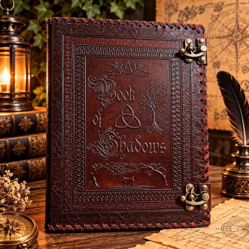 Może przedstawiać: Ciemnobrązowy dziennik w sk&oacute;rzanej oprawie o fakturowanej powierzchni, z wytłoczonym napisem Book of Shadows, symbolem triquetra, drzewem i pentagramem. Okładka jest zabezpieczona dwoma metalowymi zapięciami w kolorze mosiądzu oraz obszytymi brzegami.