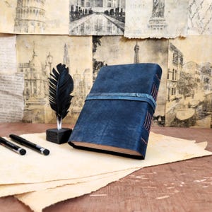 Puede incluir: Un diario de cuero azul oscuro con cierre de cinta azul y detalles de costura. El diario est&aacute; sobre una pila de papel pergamino con una pluma de pluma negra y dos bol&iacute;grafos negros.