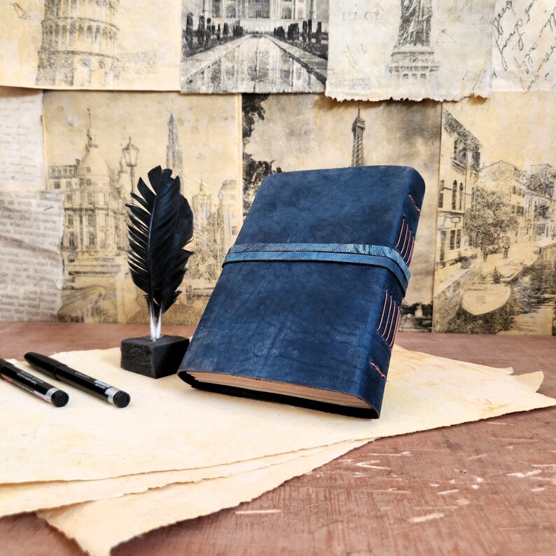 Puede incluir: Un diario de cuero azul oscuro con cierre de cinta azul y detalles de costura. El diario est&aacute; sobre una pila de papel pergamino con una pluma de pluma negra y dos bol&iacute;grafos negros.