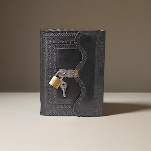 Könnte beinhalten: Ein schwarzes Lederjournal mit einem komplizierten geprägten Design und einem Schloss mit Schlüssel.