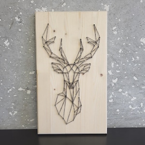 Peut inclure: Une tête de cerf géométrique réalisée en art de la ficelle sur une planche de bois. La tête de cerf est faite de ficelle noire enroulée autour de clous enfoncés dans le bois.